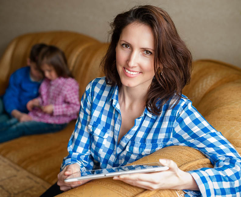 Woman smiling while holding a tablet.  Children playings in the background.